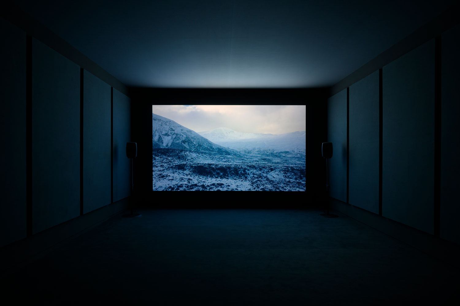 Dark room with large screen displaying a snowy mountain landscape, minimal lighting, and side speakers.