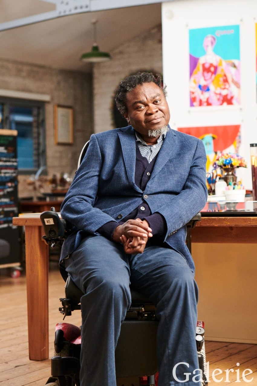 Man seated in a studio, surrounded by colorful artwork and various art supplies, wearing a blue suit and smiling gently.