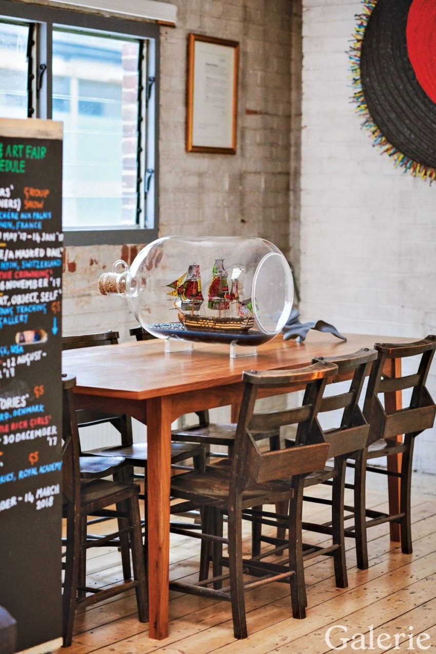 Table with chairs, a ship in a bottle centerpiece, wall art, and a poster stand in a well-lit room.