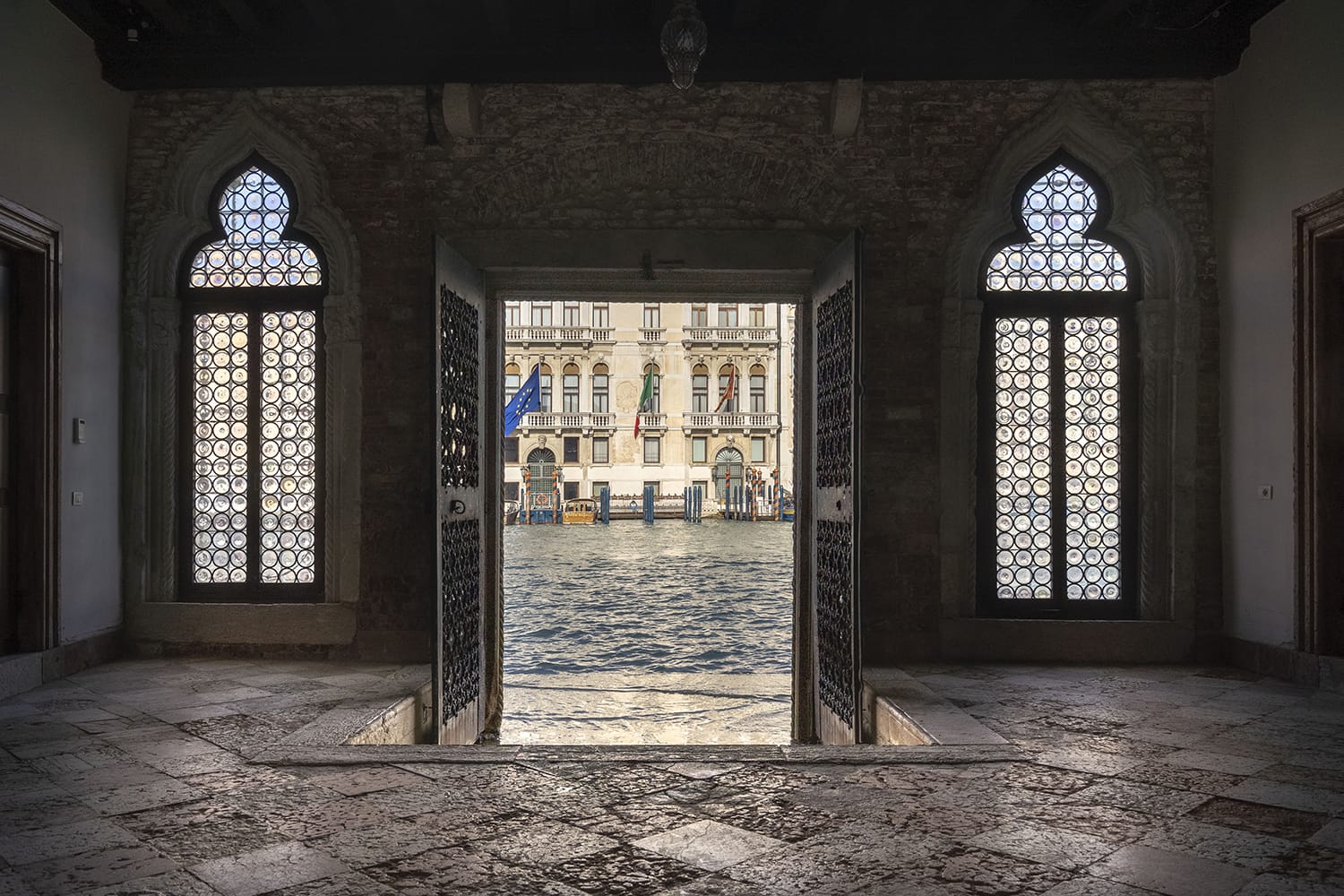 View of canal through ornate windows and open doors in a historical building, showcasing a cityscape across the water.
