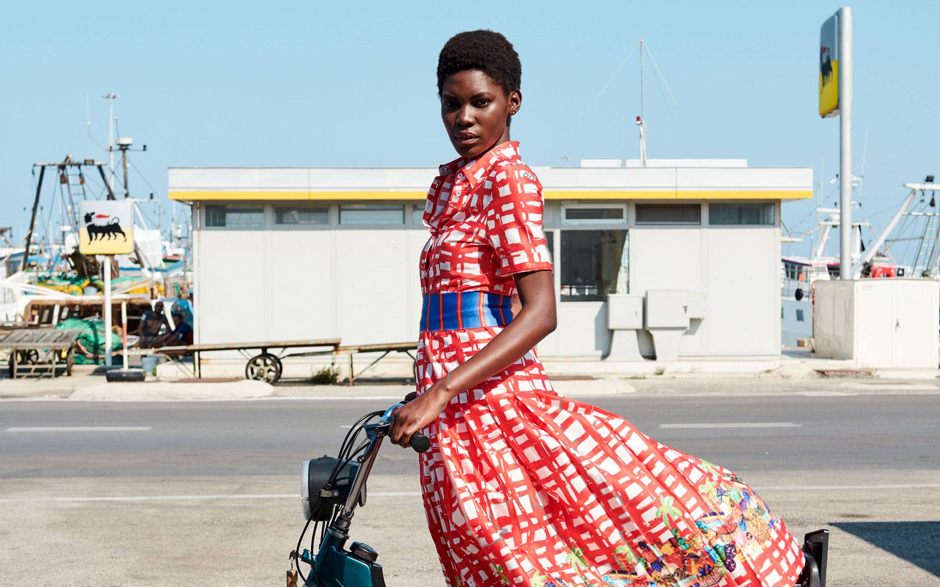 Woman in red and white checkered dress stands with bicycle near a harbor, sunny day backdrop.