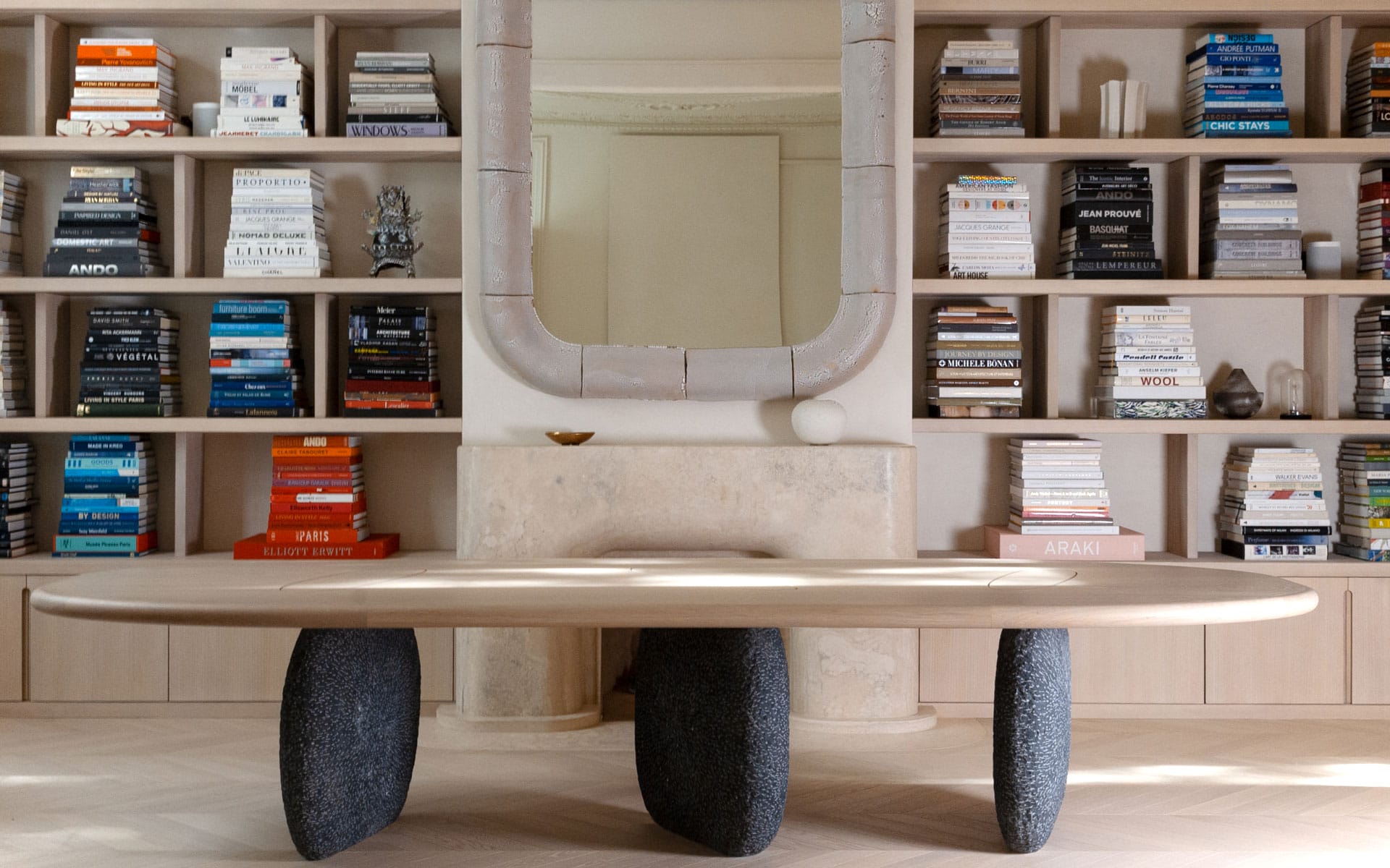 Modern wooden table with stone legs in front of a bookshelf filled with various books and a decorative mirror above.