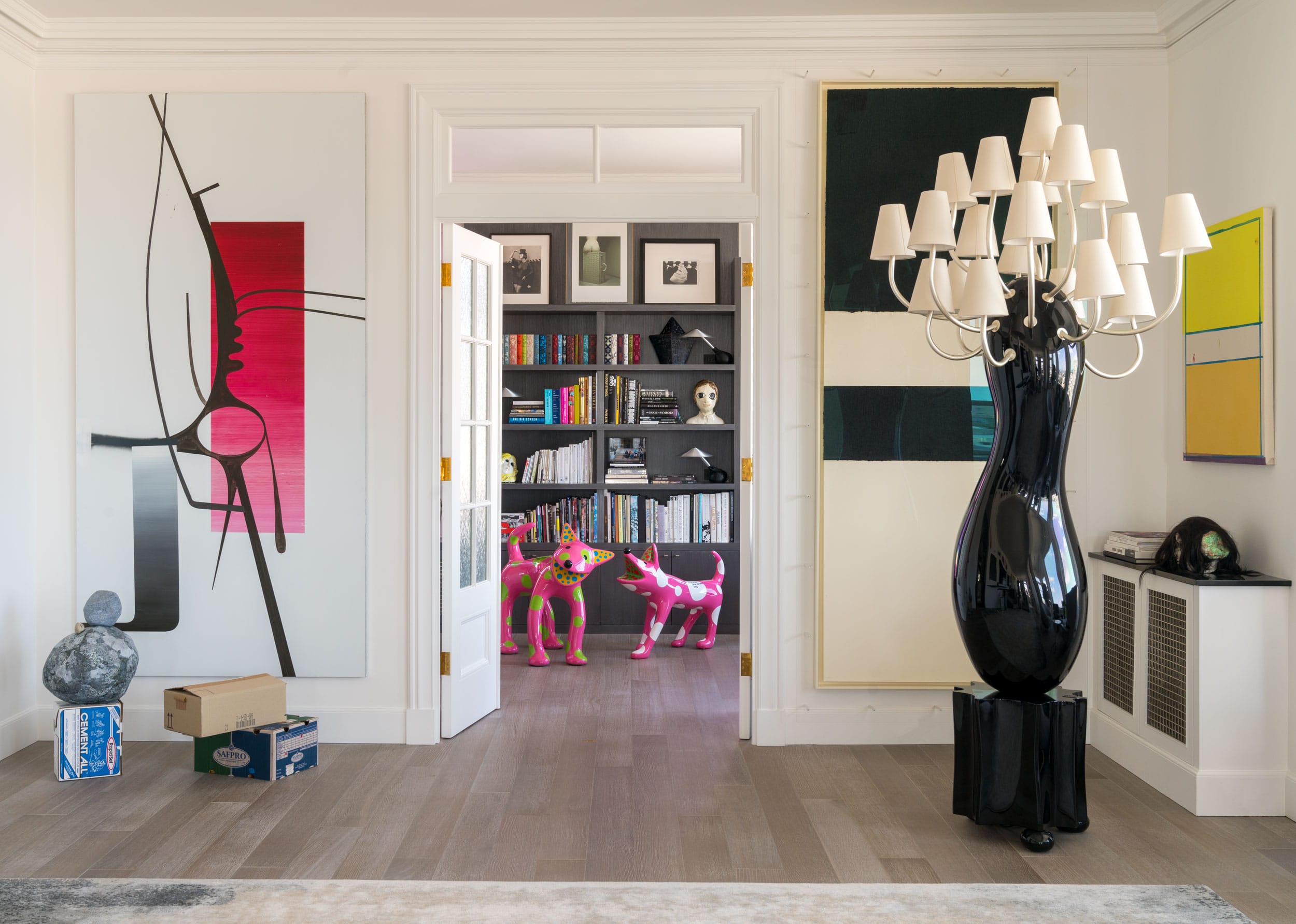 Modern living room with abstract art, large black sculpture, open doors to a bookcase, and colorful dog statues in the background.