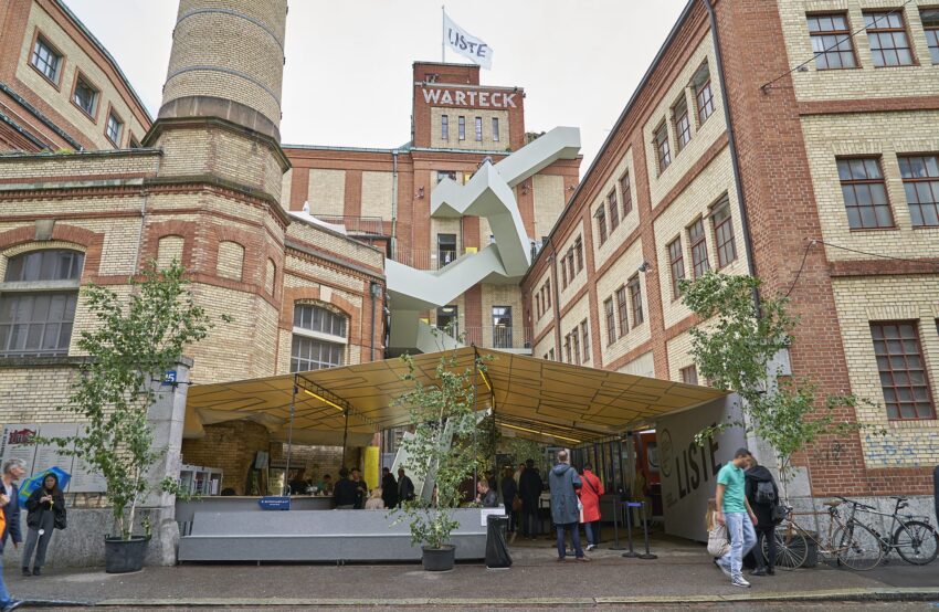 People walking under a yellow canopy in an old brick building courtyard with modern sculpture in Basel.