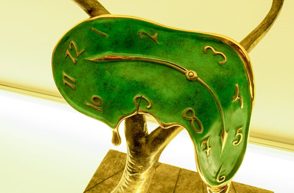 Surreal bronze sculpture of a melting green clock draped over a tree branch on a lighted surface.