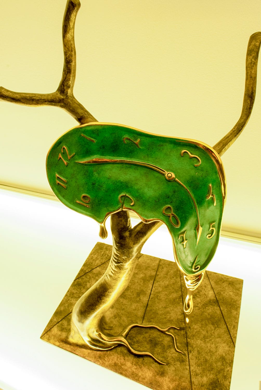 Surreal bronze sculpture of a melting green clock draped over a tree branch on a lighted surface.