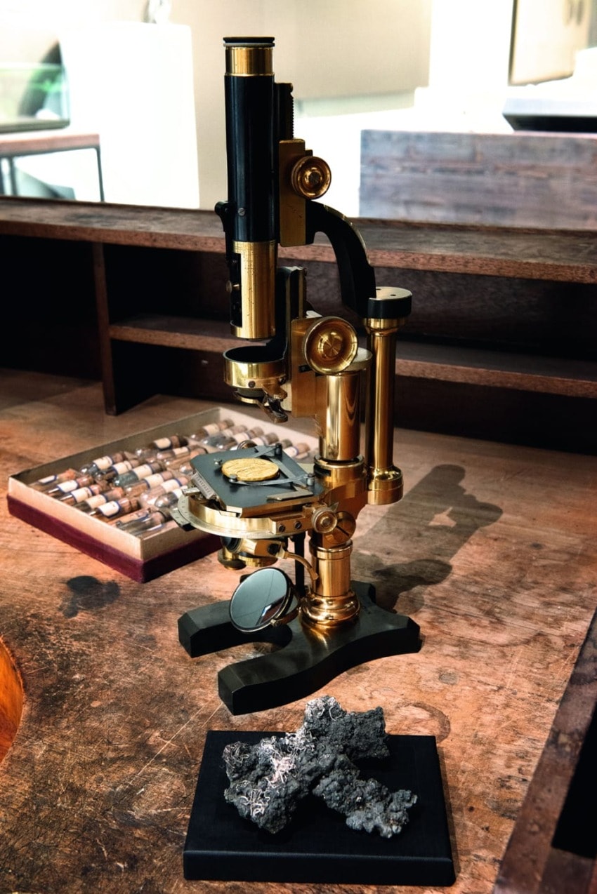 Vintage brass microscope on a wooden desk with a rock sample and a background case of small specimens.