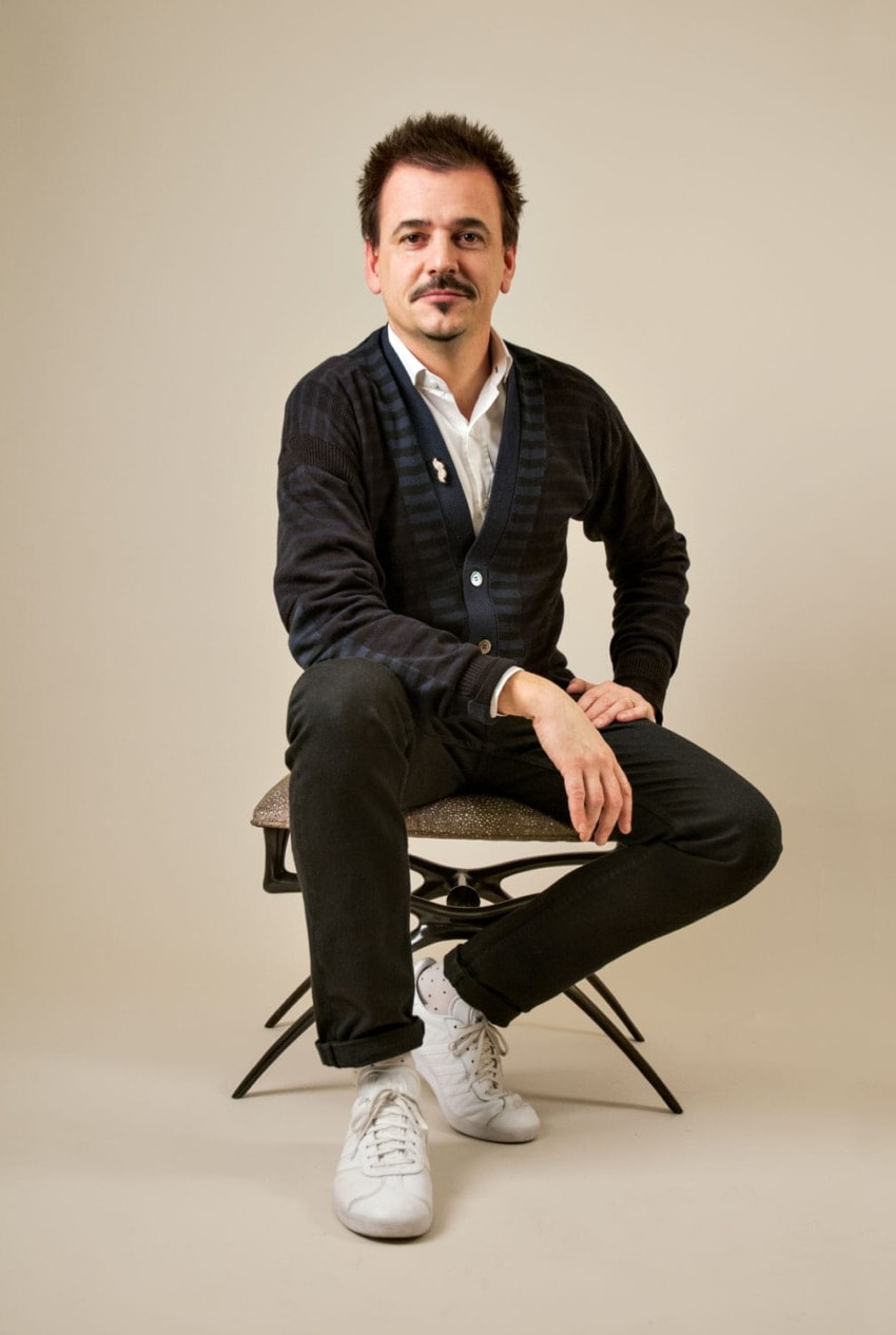 Man sitting on a chair wearing a dark cardigan, black pants, and white sneakers against a neutral background.