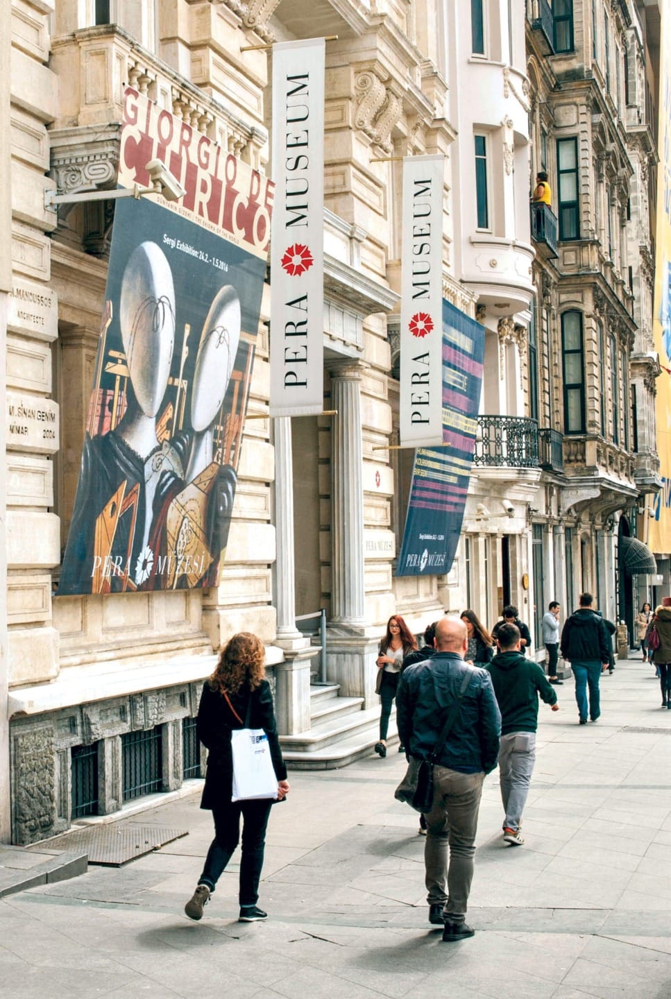 People walking outside a museum building with banners and large posters, including one featuring Giorgio de Chirico's artwork.