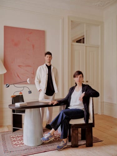 Two people in a stylish room with modern decor, one seated at a table and the other standing, with a large abstract painting.