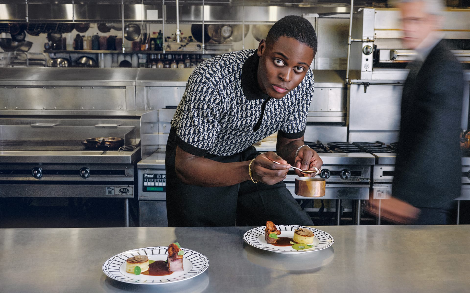 Chef plating dishes in a professional kitchen with a blurred motion figure in the background.