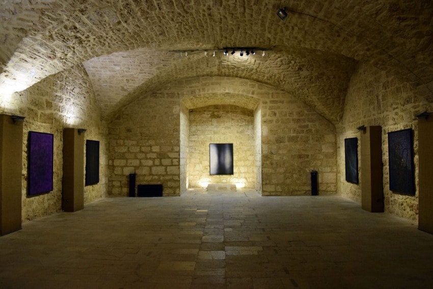 Stone-walled gallery with vaulted ceiling, dimly lit, featuring abstract paintings on the walls.