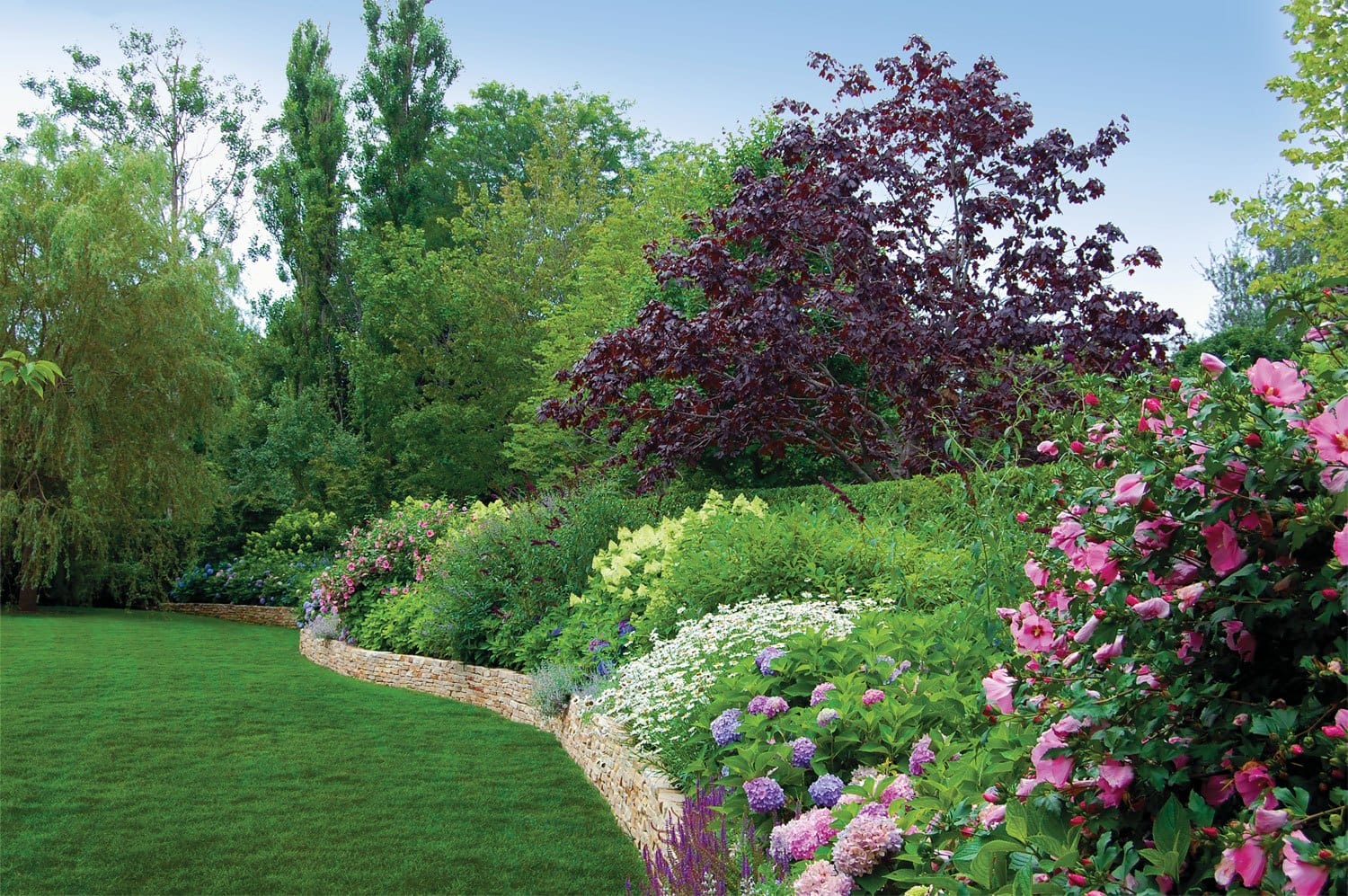 Colorful garden with blooming flowers, lush green grass, and trees under a clear sky.