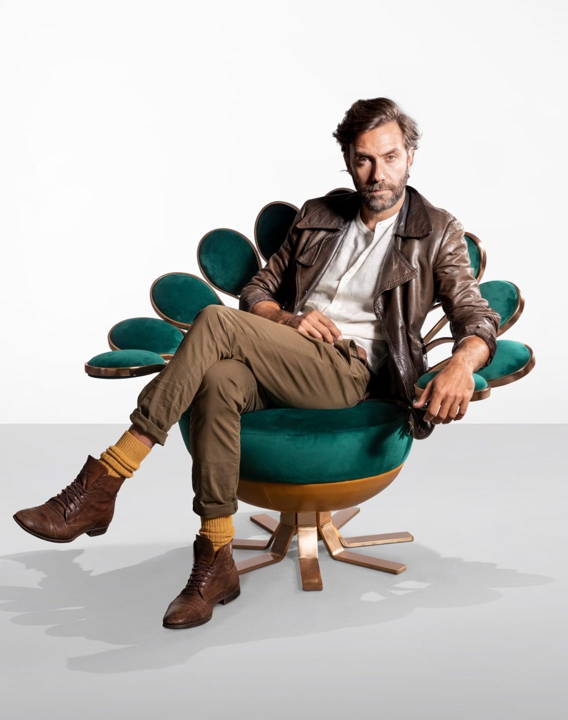 Man sitting confidently in a stylish green chair, wearing a brown leather jacket, brown boots, and yellow socks.