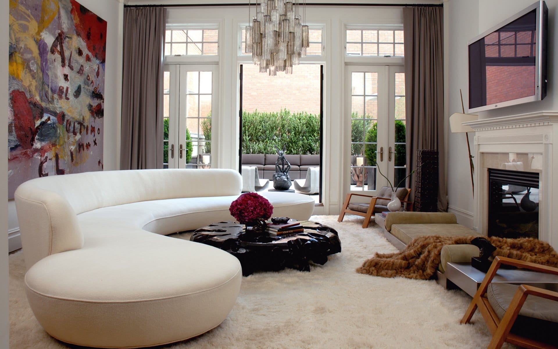 Modern living room with curved white sofa, abstract art, chandelier, and French doors leading to a patio with greenery.