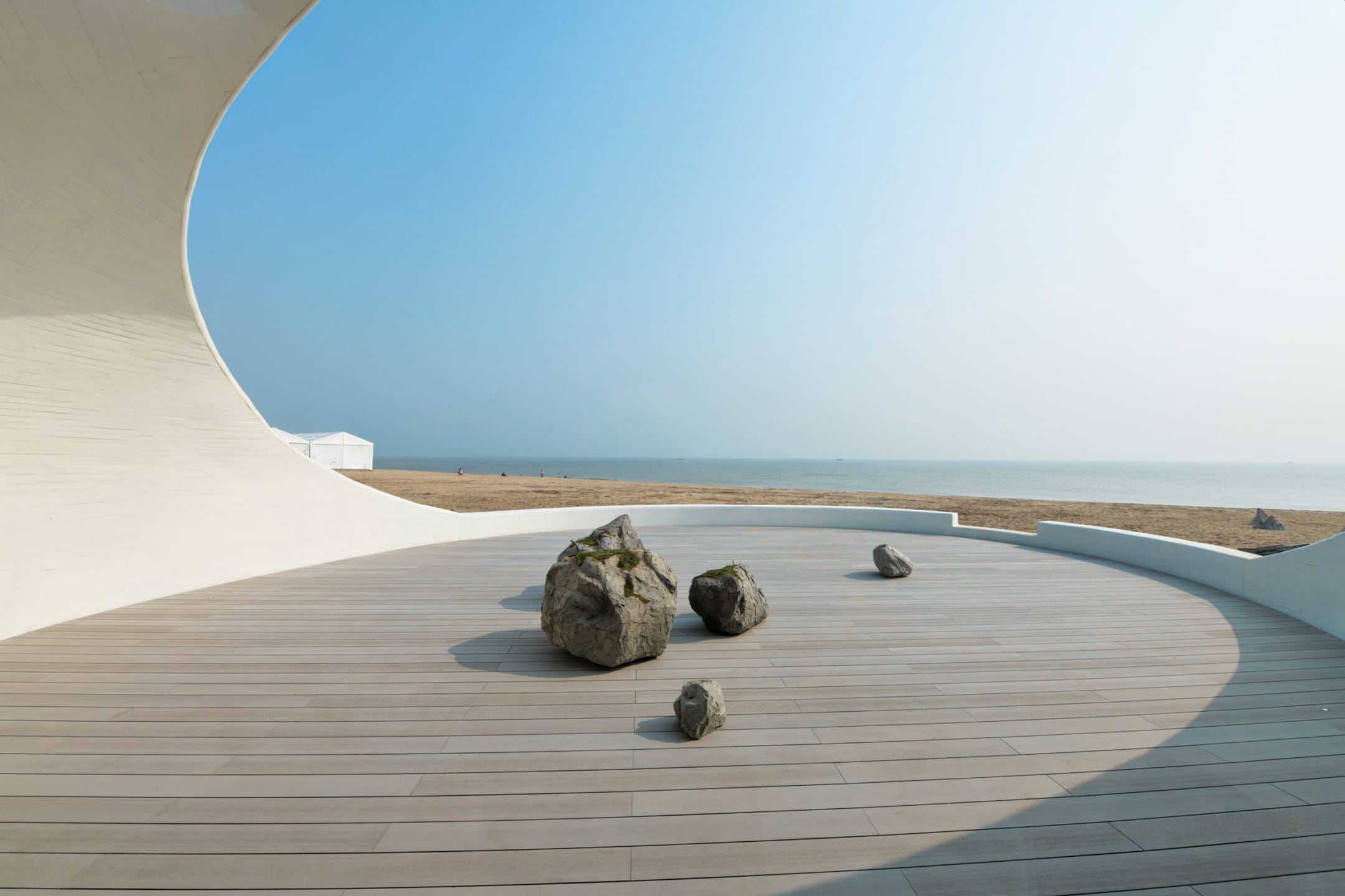 Curved modern architecture with rocks on a wooden deck overlooking a sandy beach and clear blue sky.