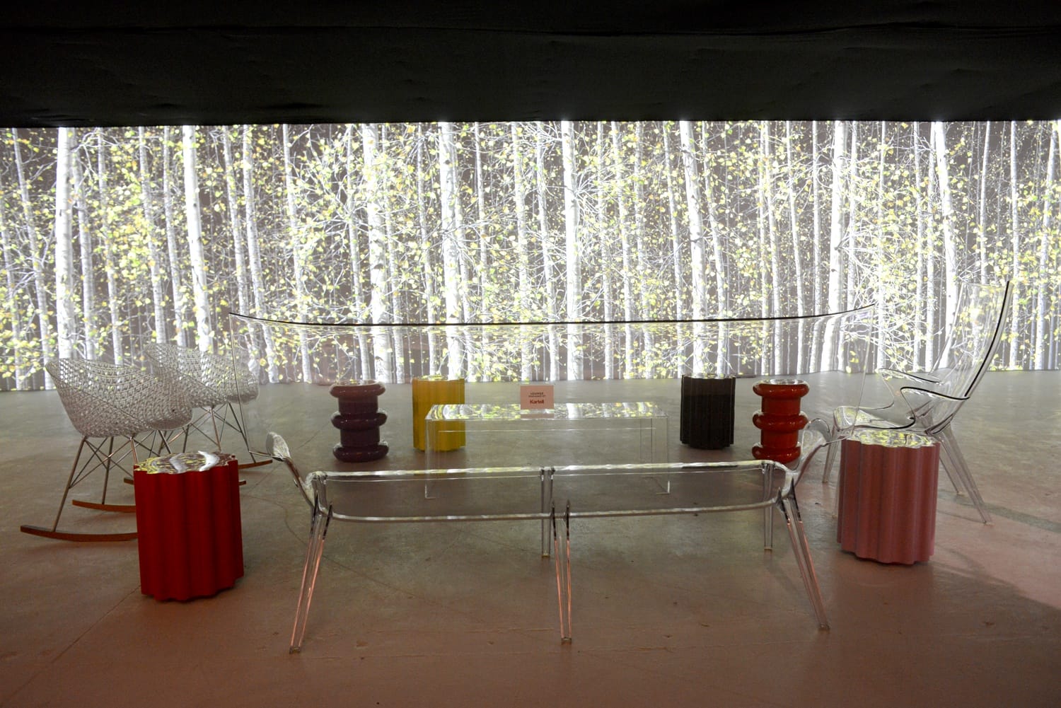 Modern art installation with transparent chairs and tables against a backdrop of tall, slender birch trees.