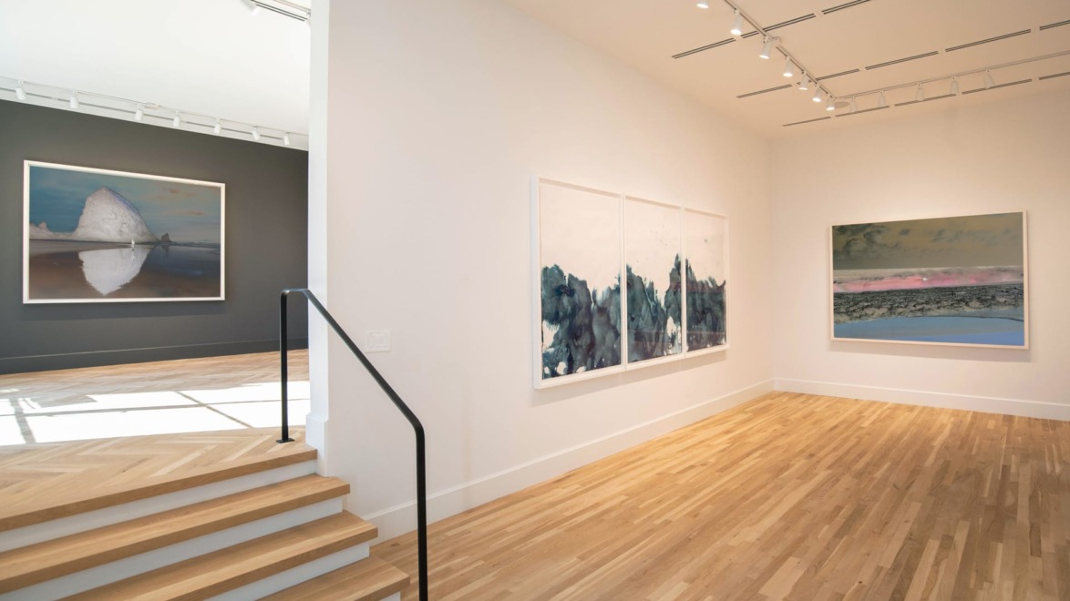 Modern art gallery interior with wooden floors, large landscape paintings on white walls, and natural light illuminating the space.