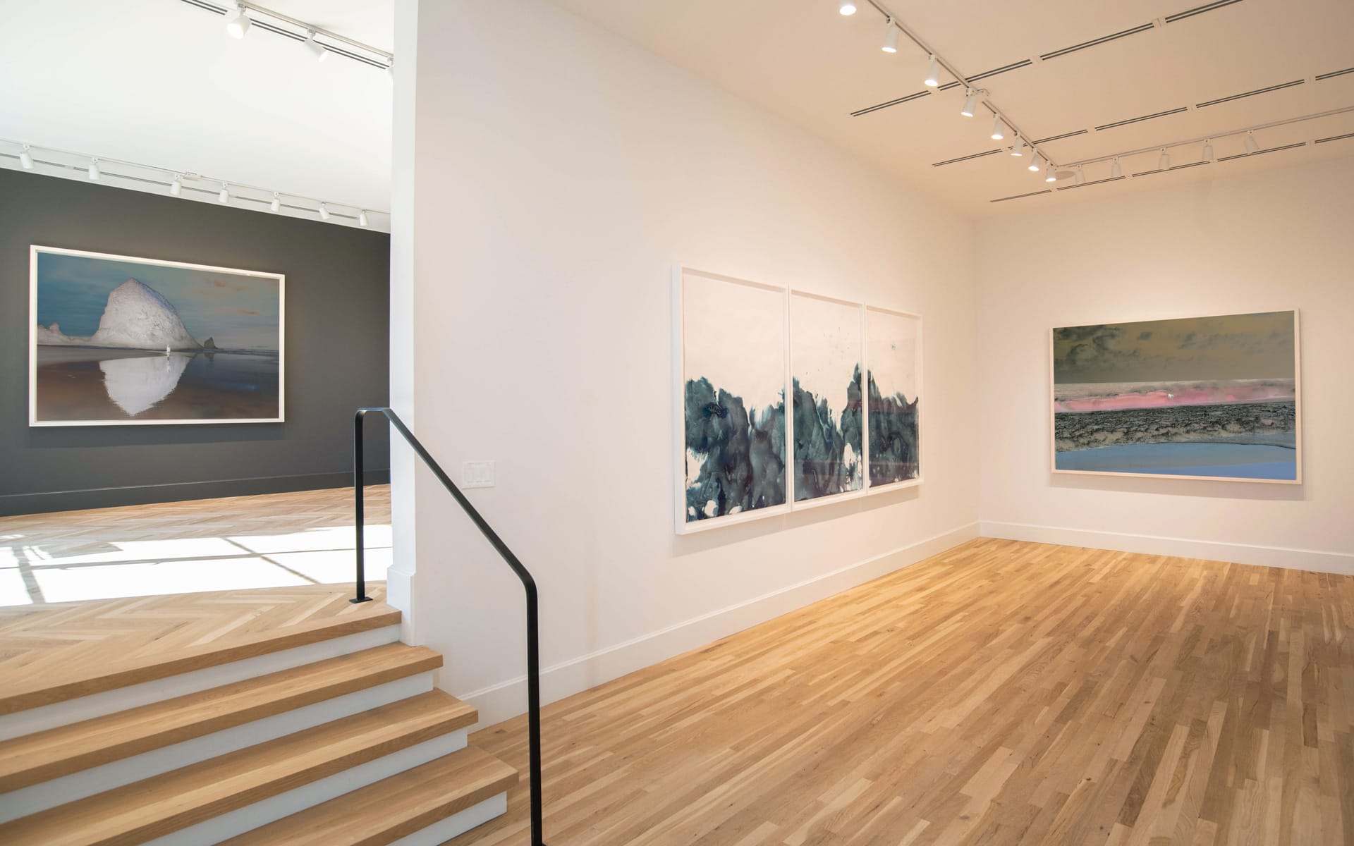 Modern art gallery interior with wooden floors, large landscape paintings on white walls, and natural light illuminating the space.