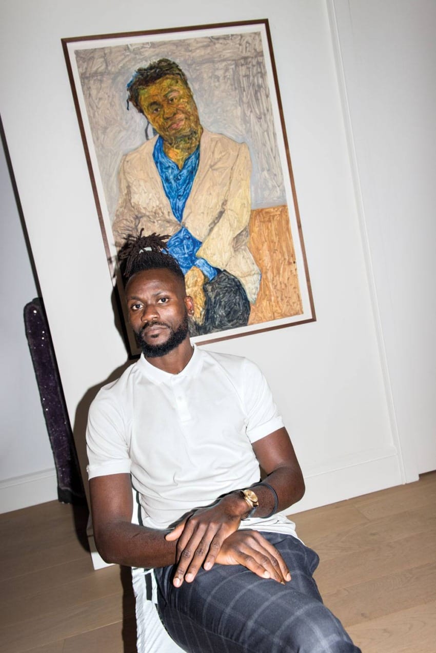 Person sitting in front of a colorful portrait painting, wearing a white shirt and dark trousers, with a contemplative expression.