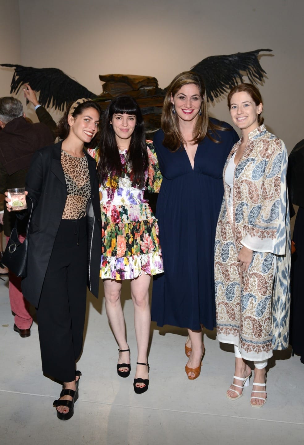 Four women smiling and posing together at an art gallery event, with an art piece visible in the background.