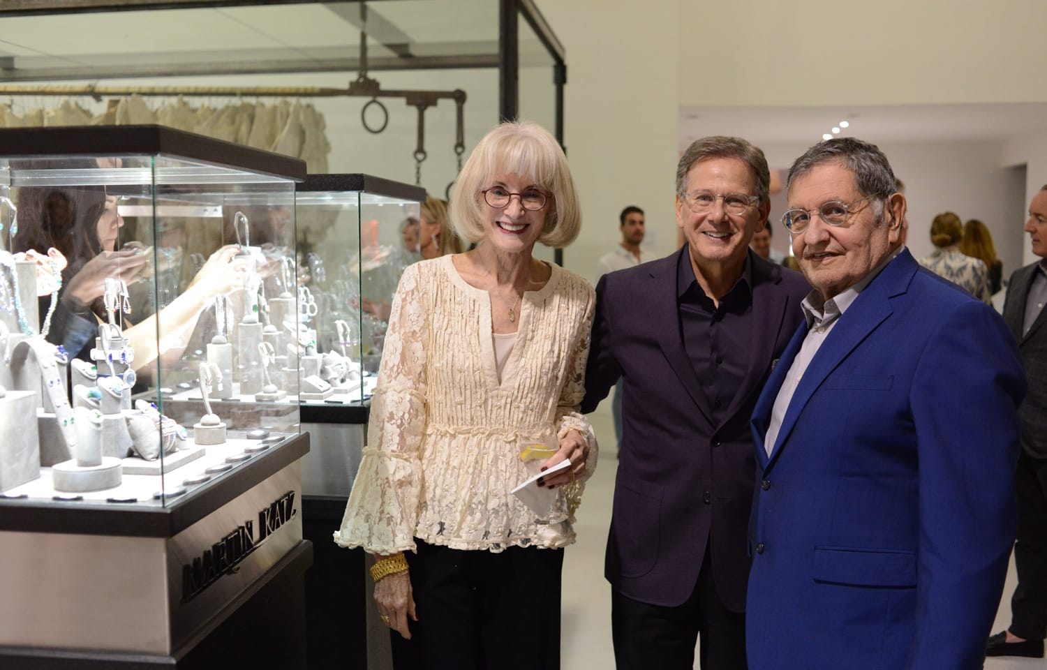Three people smiling next to a jewelry display case at an art gallery event.