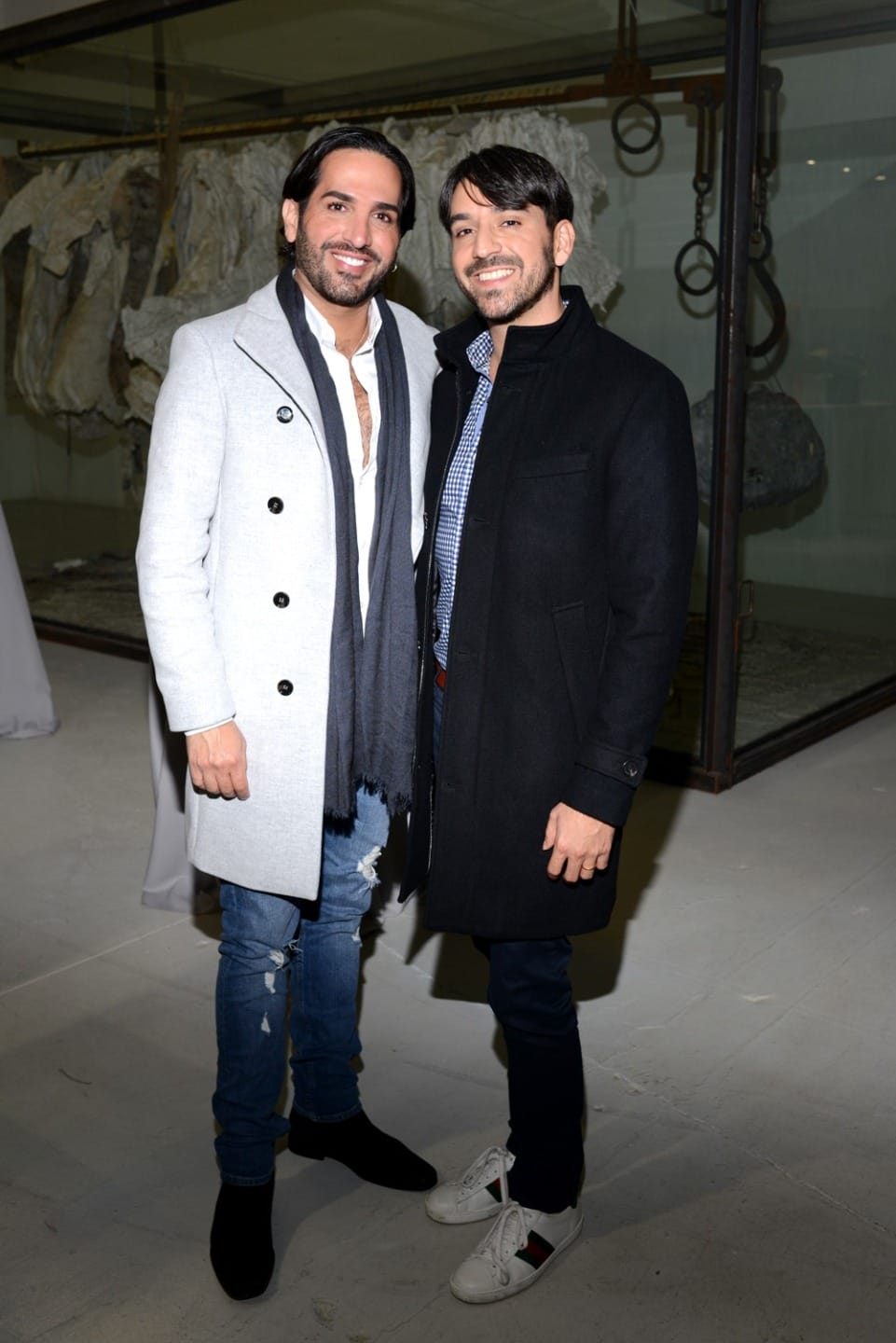 Two people in stylish winter coats pose for a photo at an indoor art exhibition.