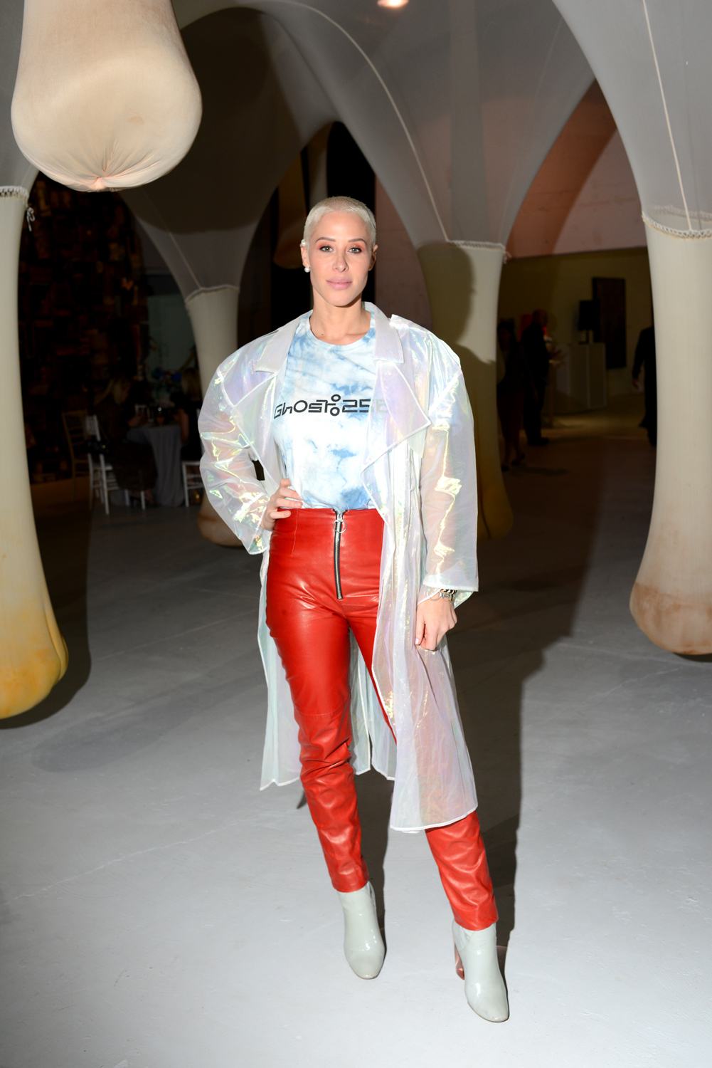 Person in a translucent coat with red pants and boots standing in an art gallery space.