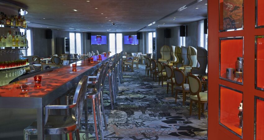 Stylish bar interior with modern decor, red accents, transparent chairs, and a long countertop under dim lighting.