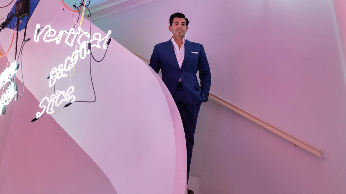 Person in a blue suit standing on a stairway with colorful neon lights in the background.