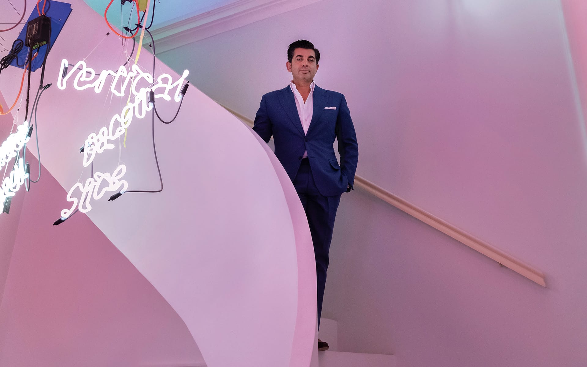 Person in a blue suit standing on a stairway with colorful neon lights in the background.