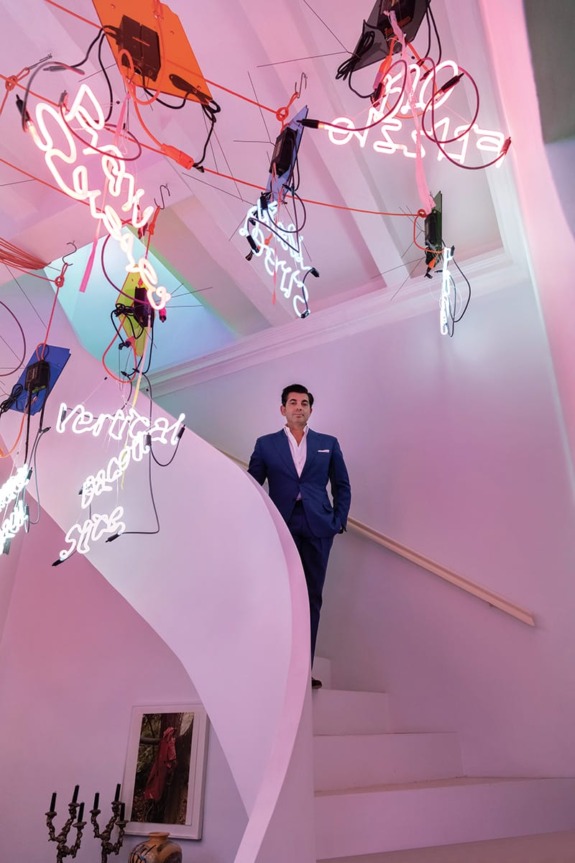 Man in a blue suit standing on a spiral staircase under colorful neon lights and art installations.