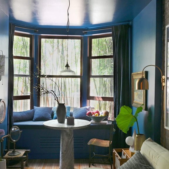 Cozy blue-painted room with bay windows, marble table, cushioned bench, and assorted decorations, including plants and artwork.