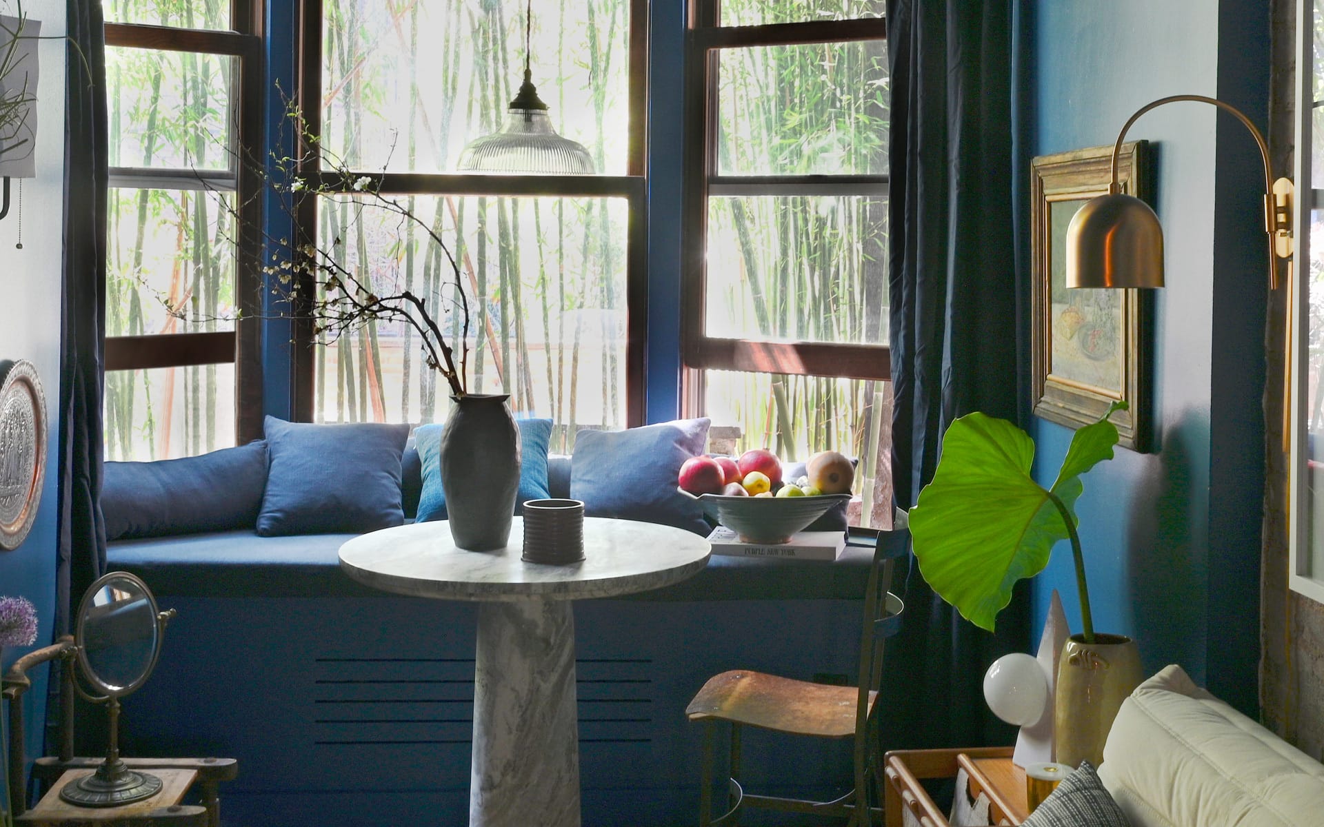 Cozy room with a round table, vase and fruit bowl by a window seat with blue cushions, overlooking bamboo trees.