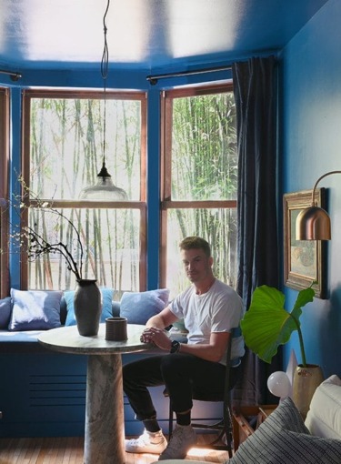 Person sitting at a round table in a blue-painted room with large windows and leafy green plants.