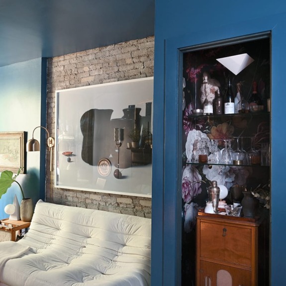 Cozy living room with a white sofa, framed artwork on brick wall, and a floral-patterned bar nook with glasses and bottles.