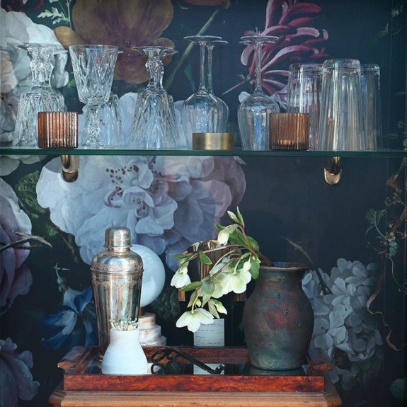 Bar shelf with glassware, cocktail shaker, potted plant, and decorative items against a floral-patterned wall.