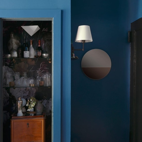 Stylish interior with blue walls, wall-mounted lamp, round mirror, small cabinet, and glassware showcase.