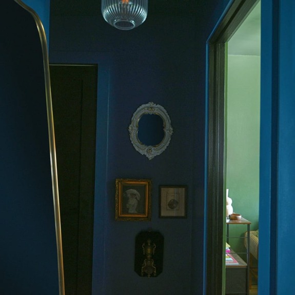 Dim hallway with dark blue walls, vintage frames, and an open door revealing a glimpse of a brightly lit green room.