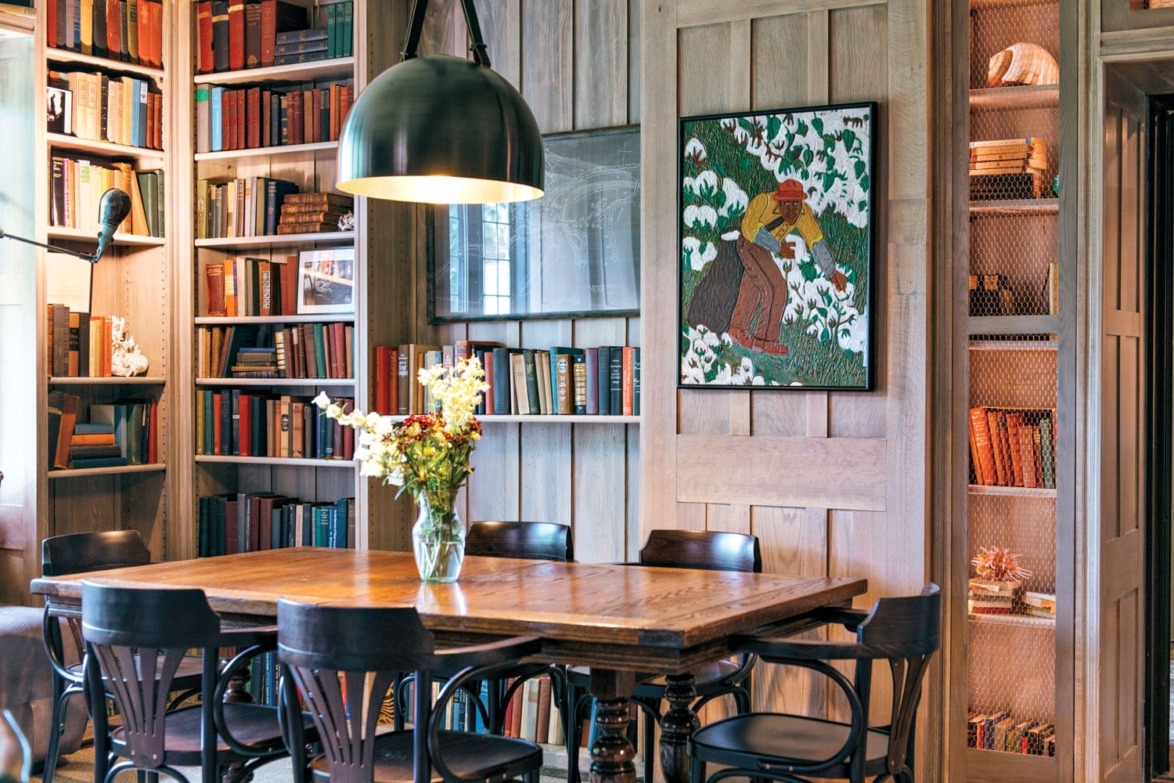 A cozy library room with a wooden table, chairs, bookshelves, a painting, and a large hanging light fixture.