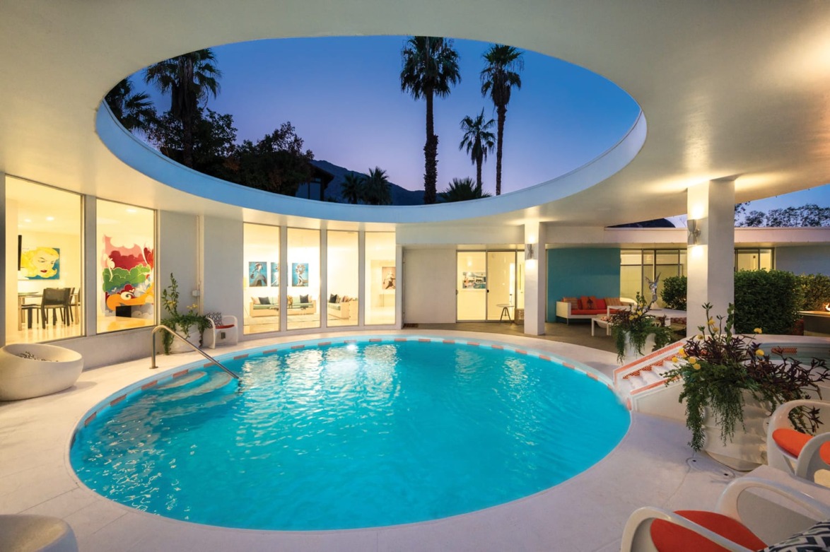 Luxurious modern house with circular indoor swimming pool, palm trees visible through open ceiling, evening setting.