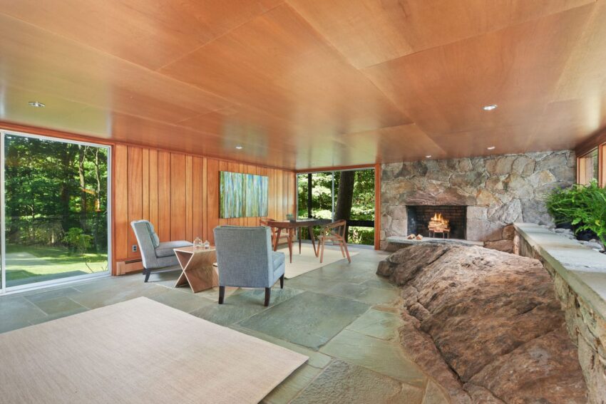 Cozy living room with wooden walls, stone fireplace, natural rock feature, modern furniture, and large windows overlooking greenery.