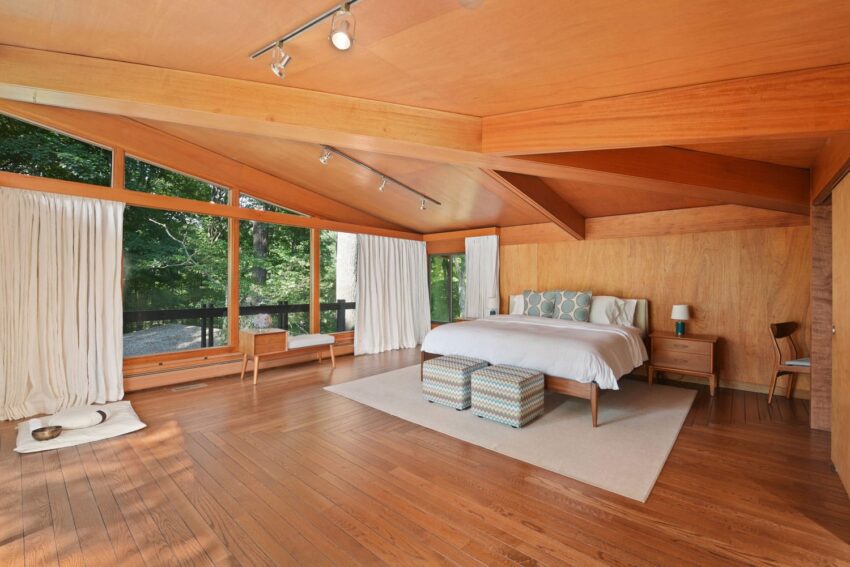 Spacious bedroom with wooden walls, large windows, a bed, and a view of greenery outside.