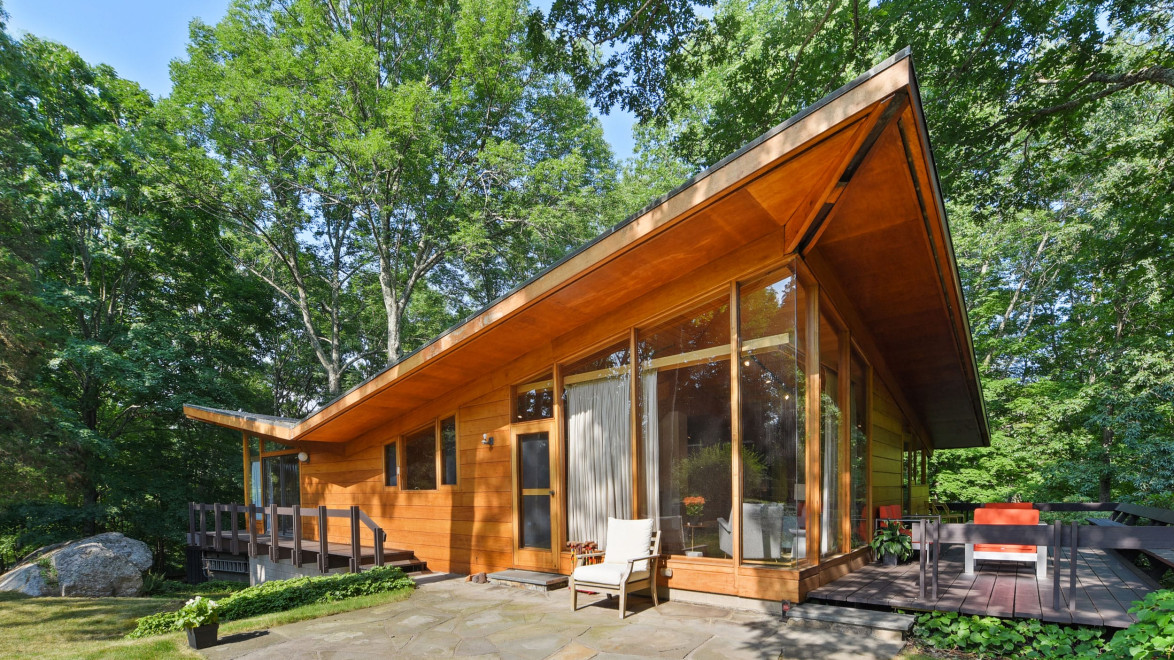 Modern wooden house with large windows surrounded by trees, featuring a stone patio and outdoor seating area.