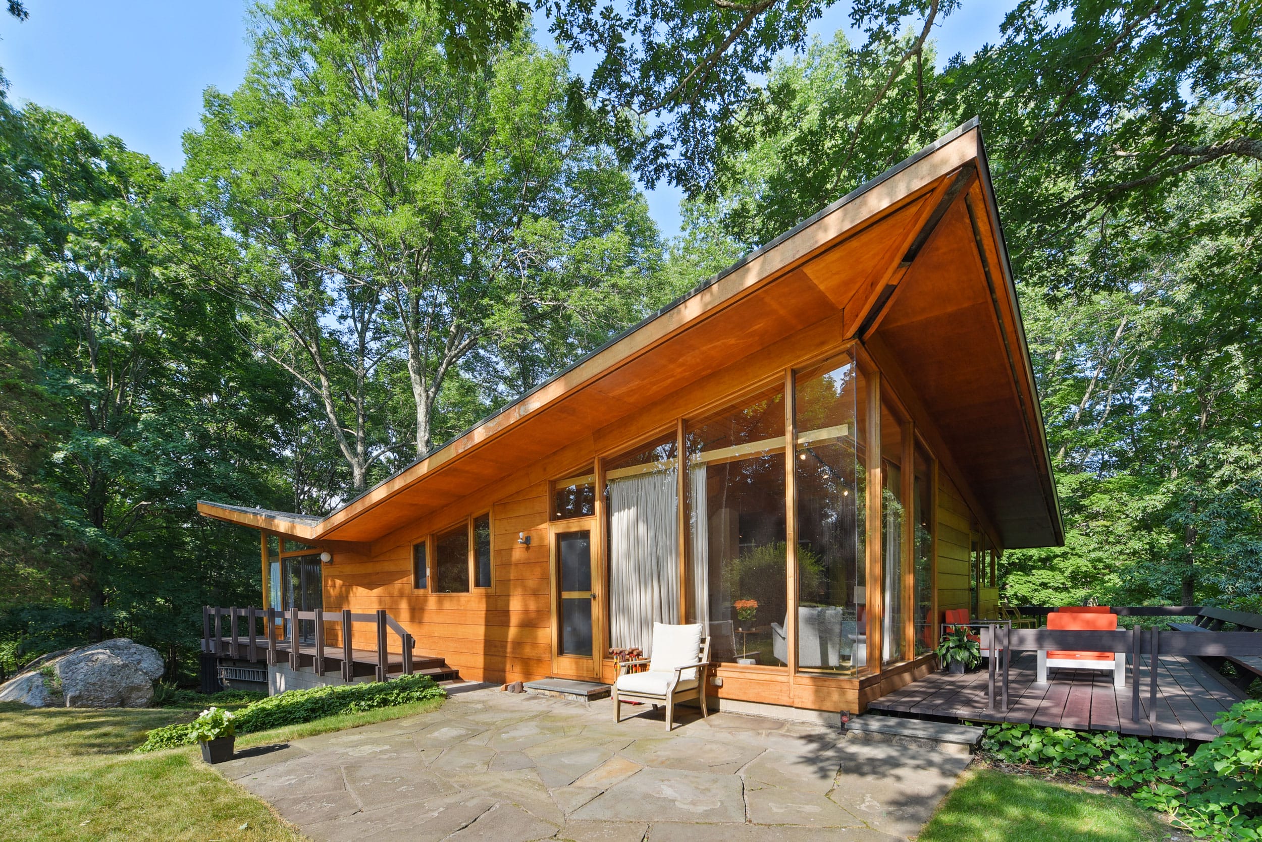 Modern wooden house with large windows surrounded by trees, featuring a stone patio and outdoor seating area.