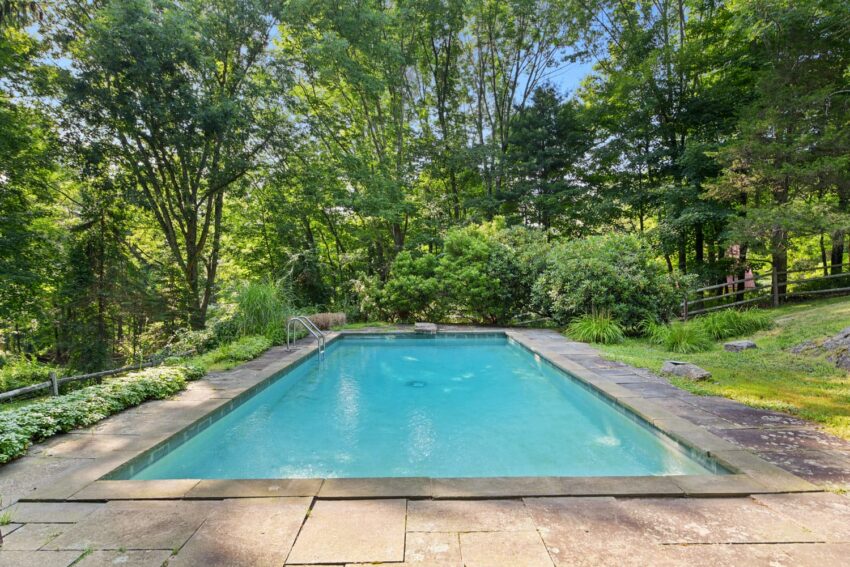 Backyard pool surrounded by lush green trees and stone patio on a sunny day.