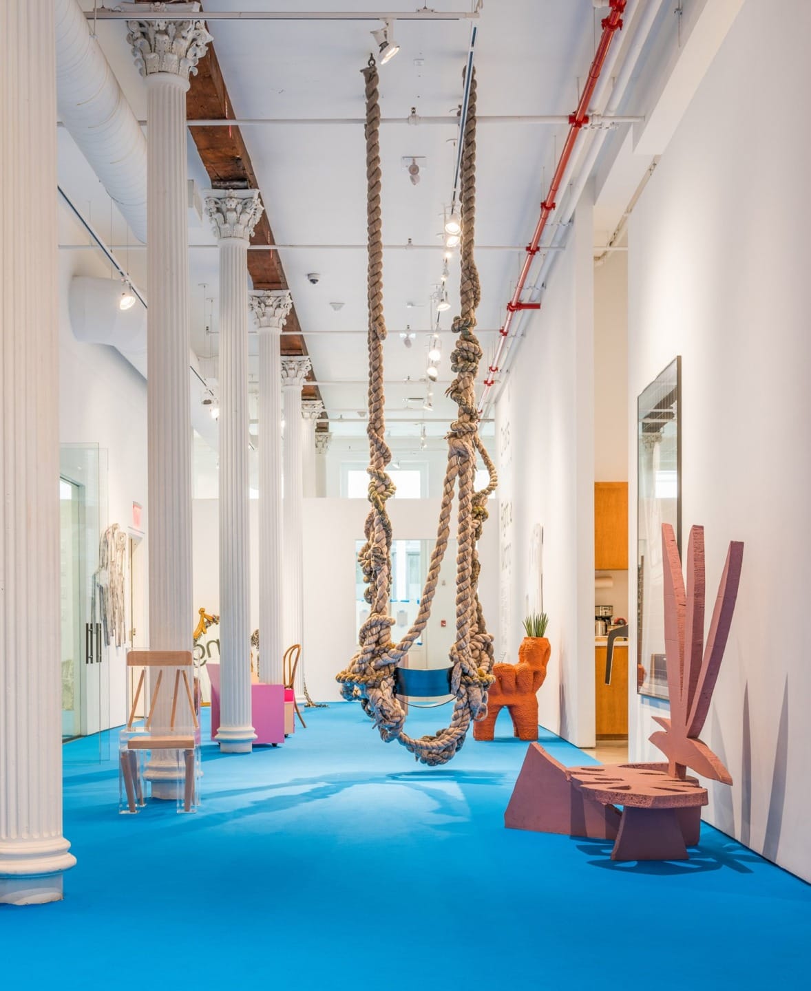 Art gallery interior with blue flooring, featuring unique sculptures, modern furniture, white columns, and rope installation.