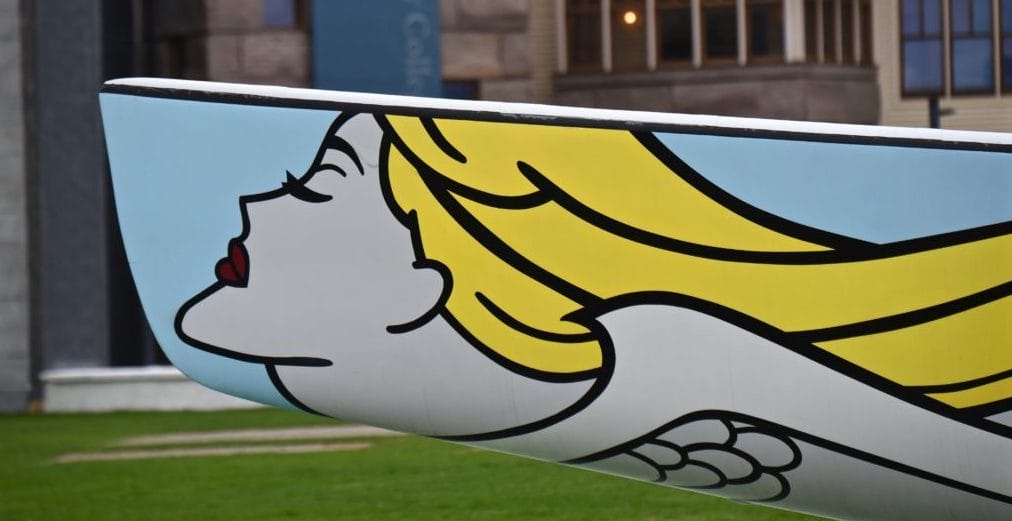 Artistic boat prow with a pop art painting of a woman's face and flowing yellow hair against a city backdrop.