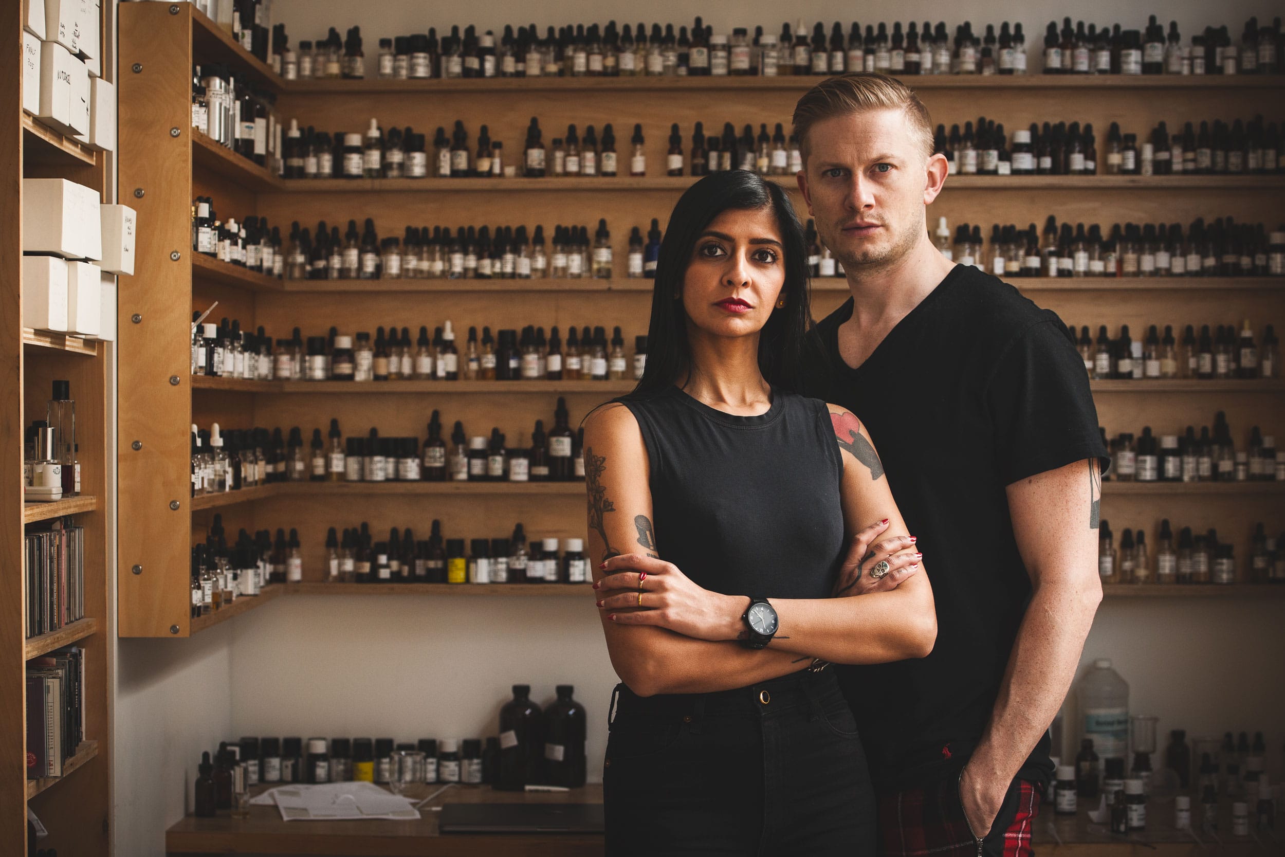 Two people standing in a room filled with shelves of bottles, looking at the camera, with a serious expression.