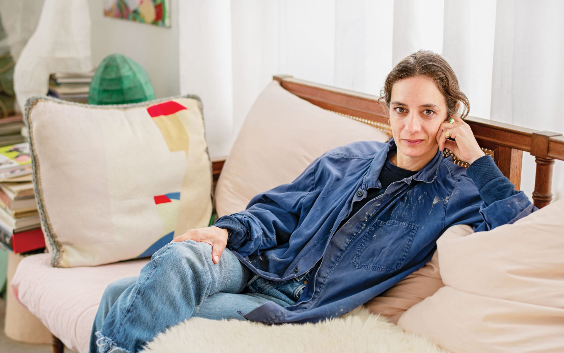 Person sitting on a couch with a relaxed pose, wearing a blue jacket and jeans, surrounded by cozy cushions and soft light.